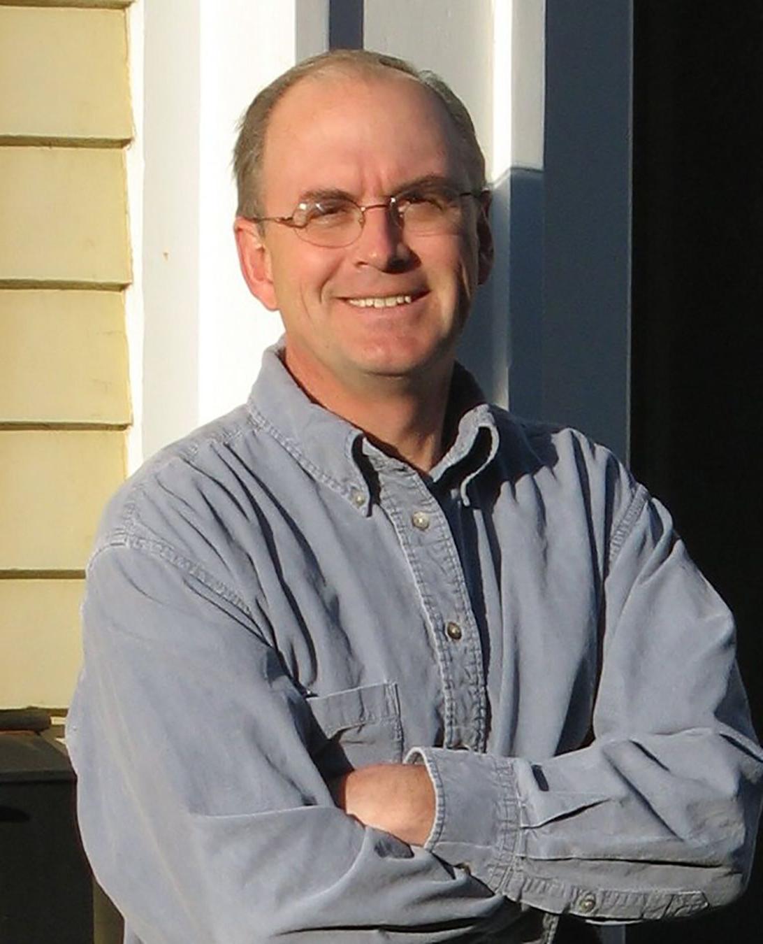 color photograph of a man wearing glasses and a gray shirt lleaning against a door frame with folded arms